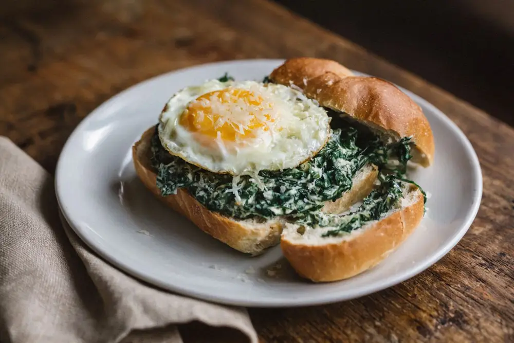 Hollowed kaiser roll filled with creamed spinach, topped with a fried egg and grated cheese on a white plate